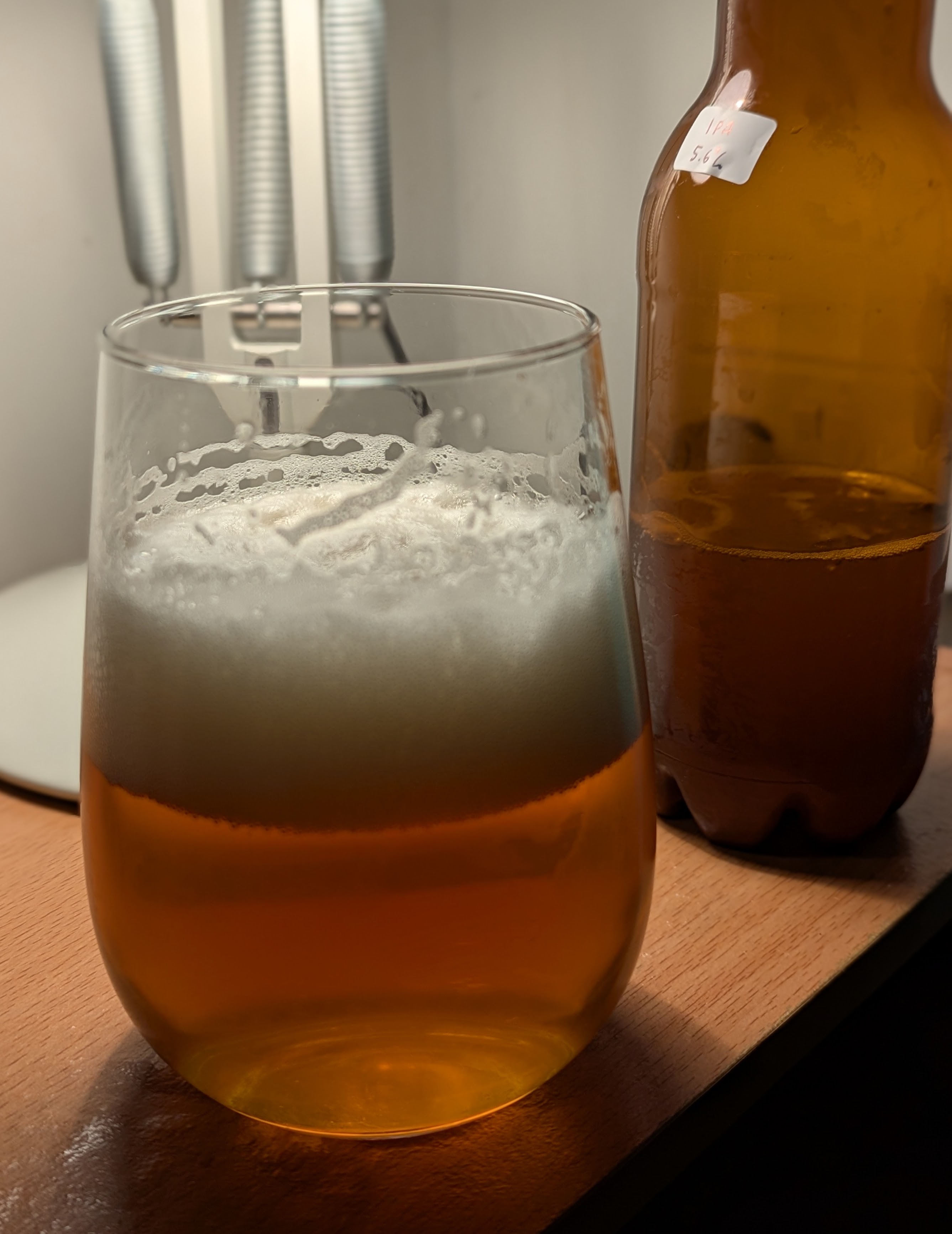 A picture of a glass of amber coloured beer with a white foamy head on a side table with a light behind the beer bottle next to it.