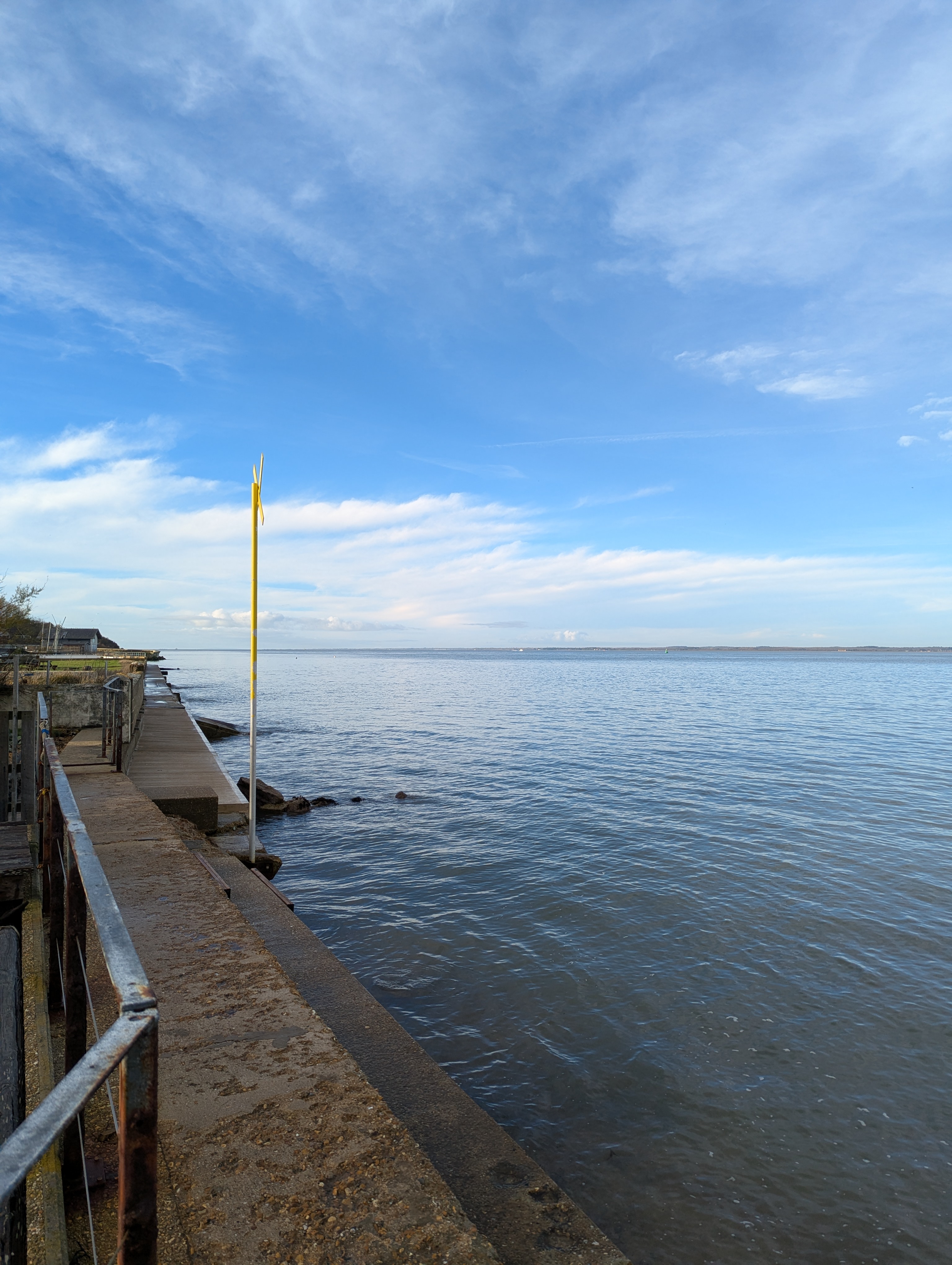 A view back over the Solent from the sea wall on Gurnard Bay of flat grey sea with blue sky and a yellow marker.